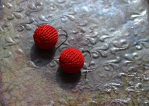 Auf einer mit Reliefdruck verzierten Kachel liegt ein Paar Ohrhänger. Diese sind aus rotem Baumwollgarn sehr fein in Form einer Kugel gehäkelt. Die Kugeln hängen an je einem Silberhaken.